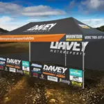 Large black Davey Motorsports tent with an orange awning, sponsor banners on a dirt lot against a desert/mountain backdrop.