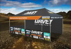 Large black Davey Motorsports tent with an orange awning, sponsor banners on a dirt lot against a desert/mountain backdrop.