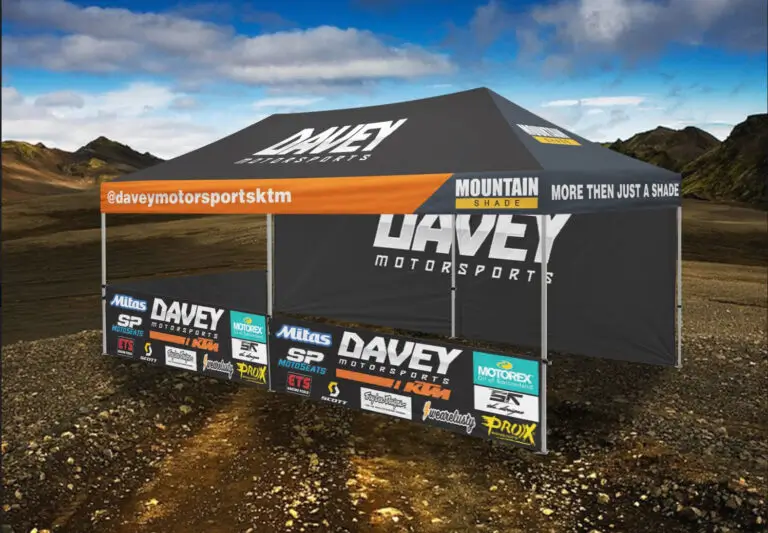 Large black Davey Motorsports tent with an orange awning, sponsor banners on a dirt lot against a desert/mountain backdrop.