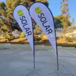 Two teardrop banners with the word SOLAR and a green-yellow logo stand in a parking lot on a sunny day, with trees in the background.
